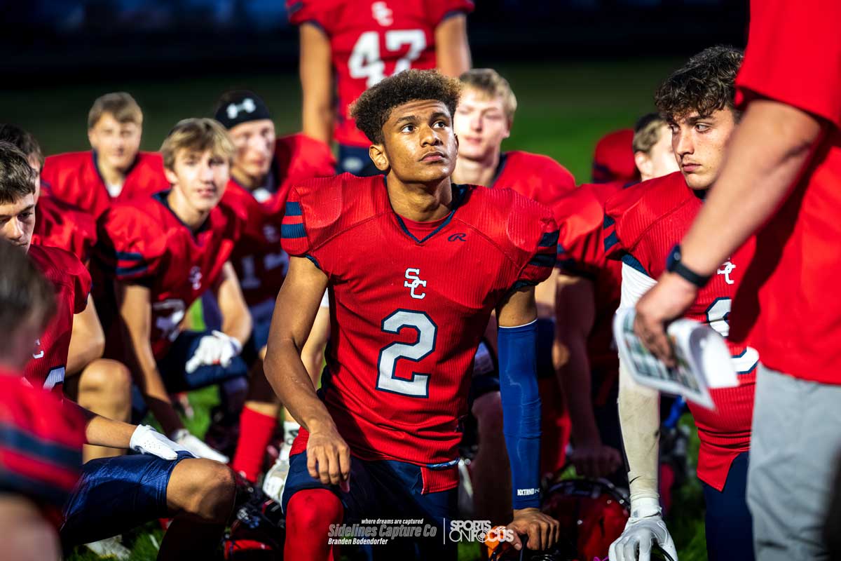 Spencer/Columbus football player kneeling with teammates during a sideline huddle under the lights